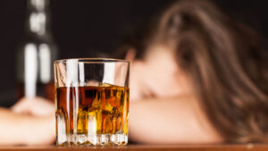 Bicchiere di whisky su un tavolo con ghiaccio, in primo piano. Sullo sfondo sfocato, una donna con la testa abbassata, simbolo degli effetti dell'alcolismo.