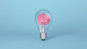 Illustrazione concettuale su sfondo azzurro che raffigura una lampadina trasparente al cui interno, al posto del filamento, è presente un cervello rosa tridimensionale. L'immagine simboleggia l’unione tra mente e tecnologia, evocando idee di innovazione e progresso scientifico nell’ambito delle neuroscienze.